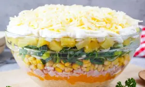 Schichtsalat mit Eisbergsalat, Mais, Schinken, Ananas, Lauch und Käse in einer Glasschüssel.