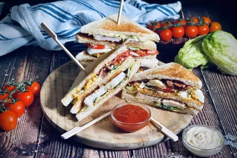 Ein gestapelter Club Sandwich mit Hühnchen, Speck, Ei und Salat, serviert mit Tomaten und Dips.