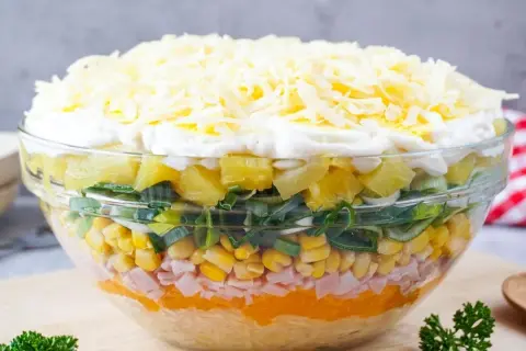 Schichtsalat mit Eisbergsalat, Mais, Schinken, Ananas, Lauch und Käse in einer Glasschüssel.