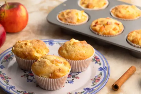 Saftige apfelmuffins ohne zucker, frisch gebacken und auf einem Teller mit Blumenmuster arrangiert. Daneben liegen Äpfel und Zimt.