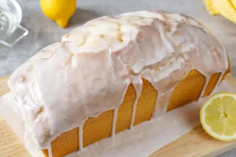 Ein saftiger Zitronenkuchen in Kastenform, überzogen mit süßem Zuckerguss. Ein Zitronenstück liegt daneben.