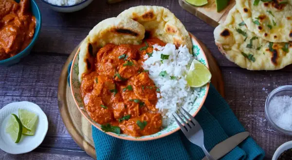 Ein Teller mit cremigem Butter Chicken, Reis und Naan-Brot. Garniert mit frischem Koriander und Limettenspalten.