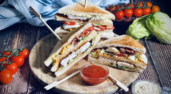 Ein gestapelter Club Sandwich mit Hühnchen, Speck, Ei und Salat, serviert mit Tomaten und Dips.