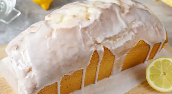 Ein saftiger Zitronenkuchen in Kastenform, überzogen mit süßem Zuckerguss. Ein Zitronenstück liegt daneben.