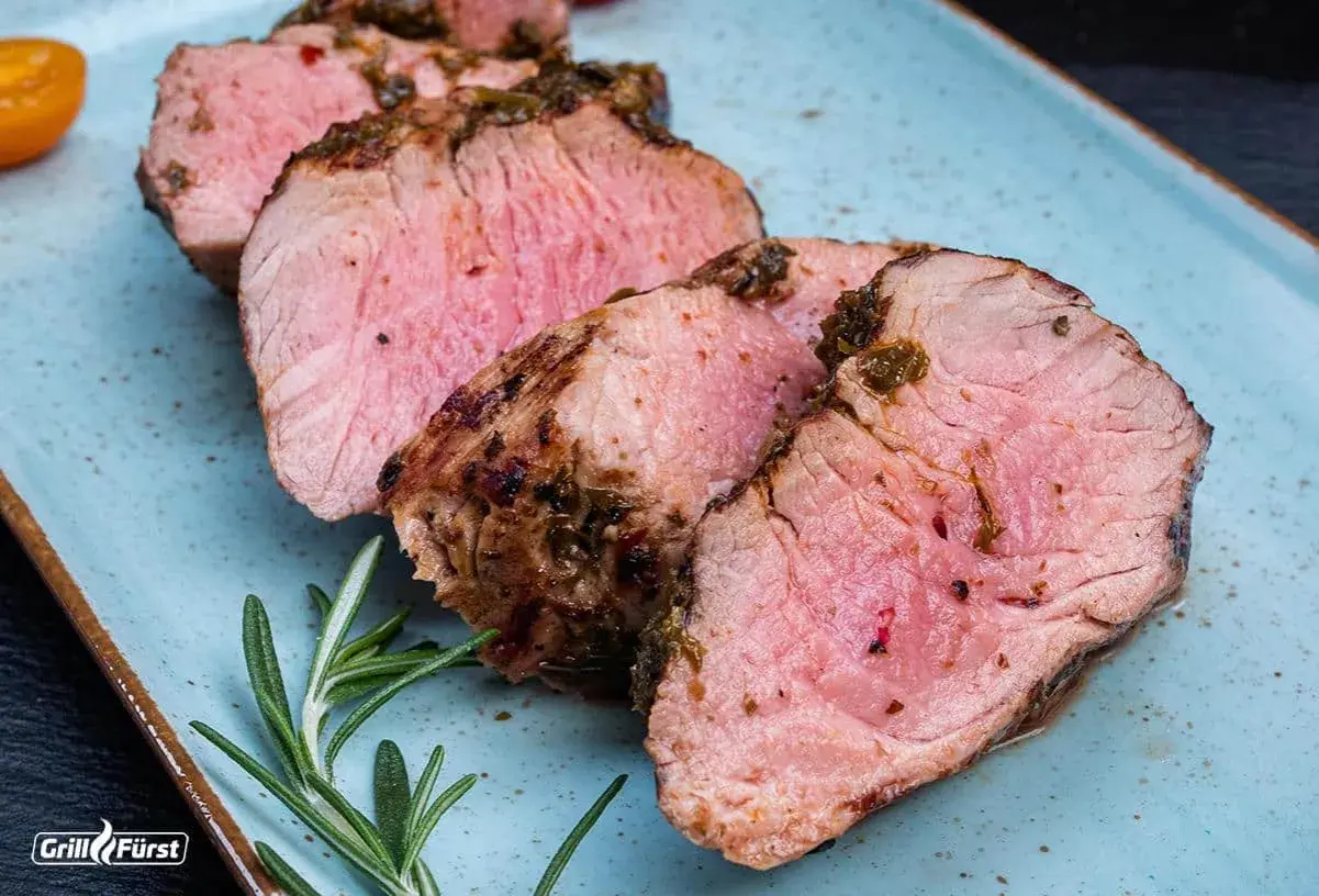 Saftiges Schweinefilet, perfekt gegrillt. Die Kerntemperatur ist entscheidend für dieses zarte Ergebnis. Rosmarin und Tomaten garnieren den Teller.