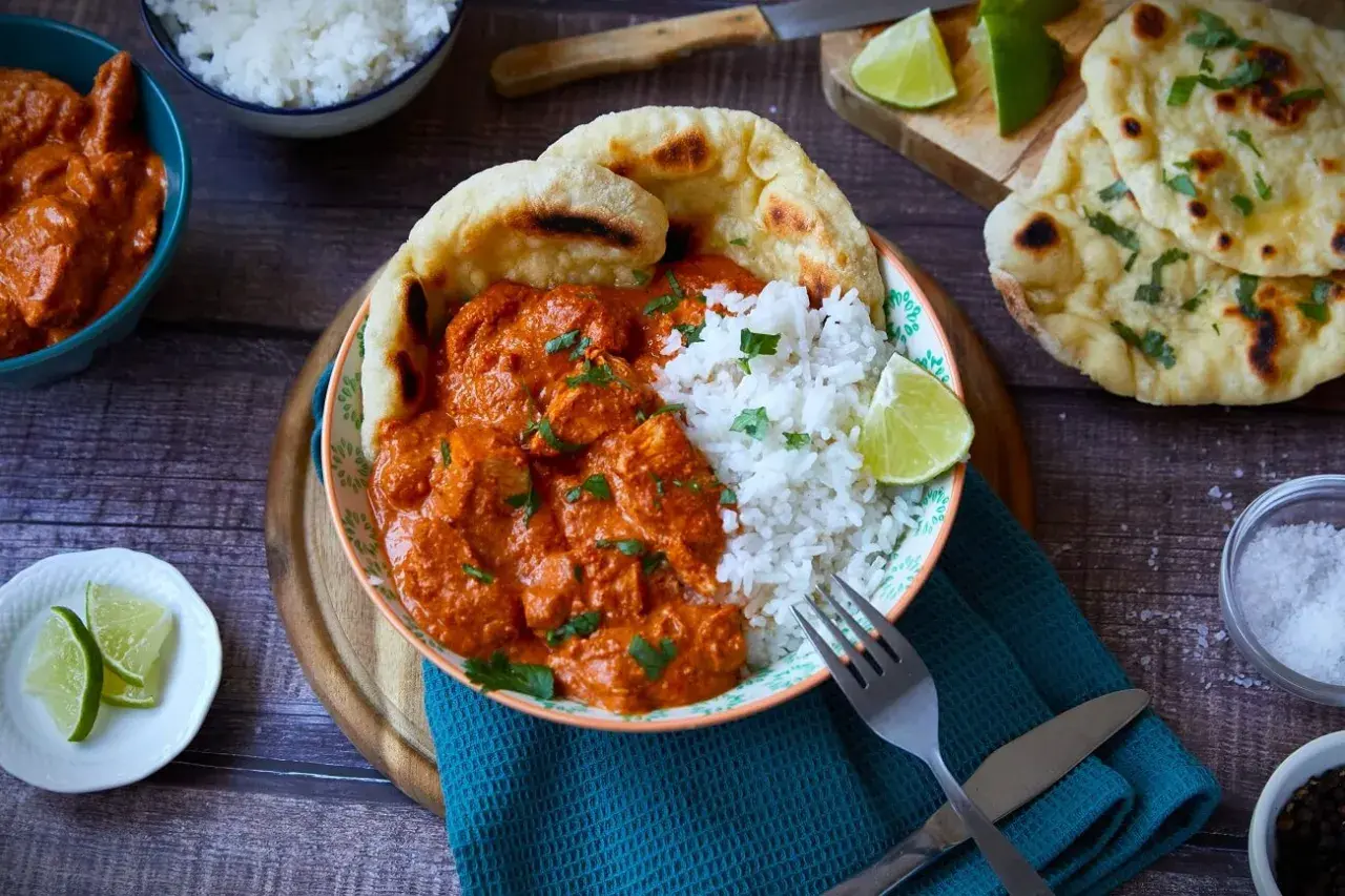 Ein Teller mit cremigem Butter Chicken, Reis und Naan-Brot. Garniert mit frischem Koriander und Limettenspalten.