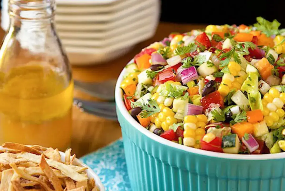 Frischer, bunter mexikanischer Salat mit Mais, Bohnen, Tomaten und Koriander in einer türkisfarbenen Schüssel, daneben eine Flasche Dressing und Tortilla-Streifen.