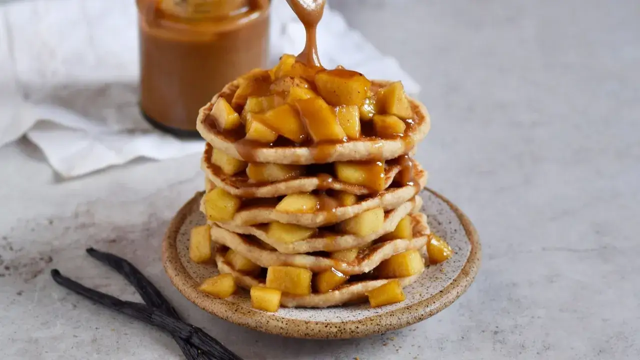 Ein Stapel fluffiger Pfannkuchen mit Hafermilch, belegt mit karamellisierten &Auml;pfeln und Sauce. Daneben liegen Vanilleschoten.
