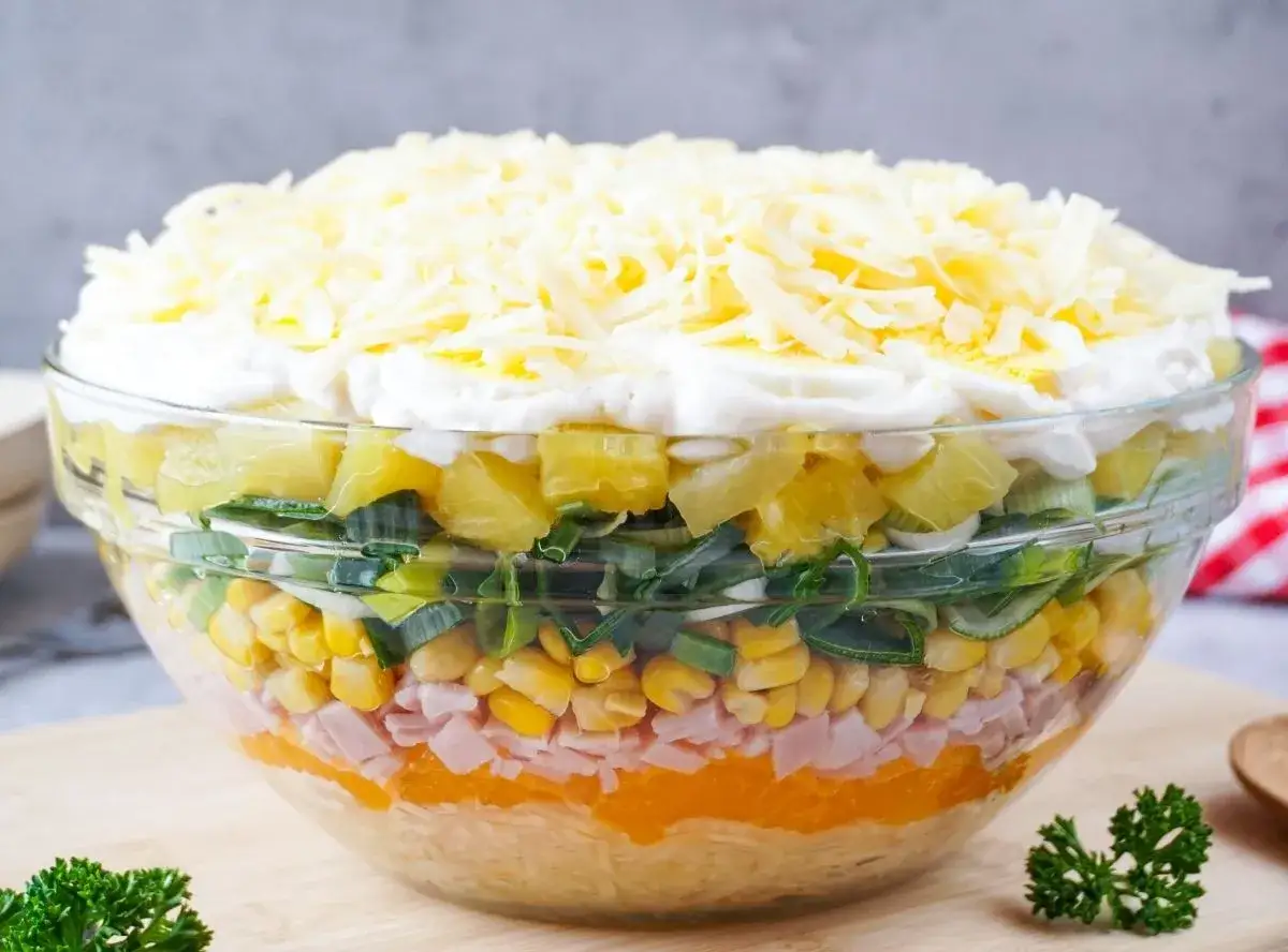 Schichtsalat mit Eisbergsalat, Mais, Schinken, Ananas, Lauch und K&auml;se in einer Glassch&uuml;ssel.