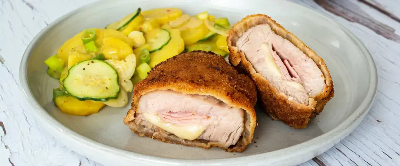Zwei goldbraune Cordon-Bleu-Portionen mit geschmolzenem K&auml;se, serviert mit einem bunten Kartoffel-Zucchini-Salat. Ein k&ouml;stliches cordon bleu rezept.