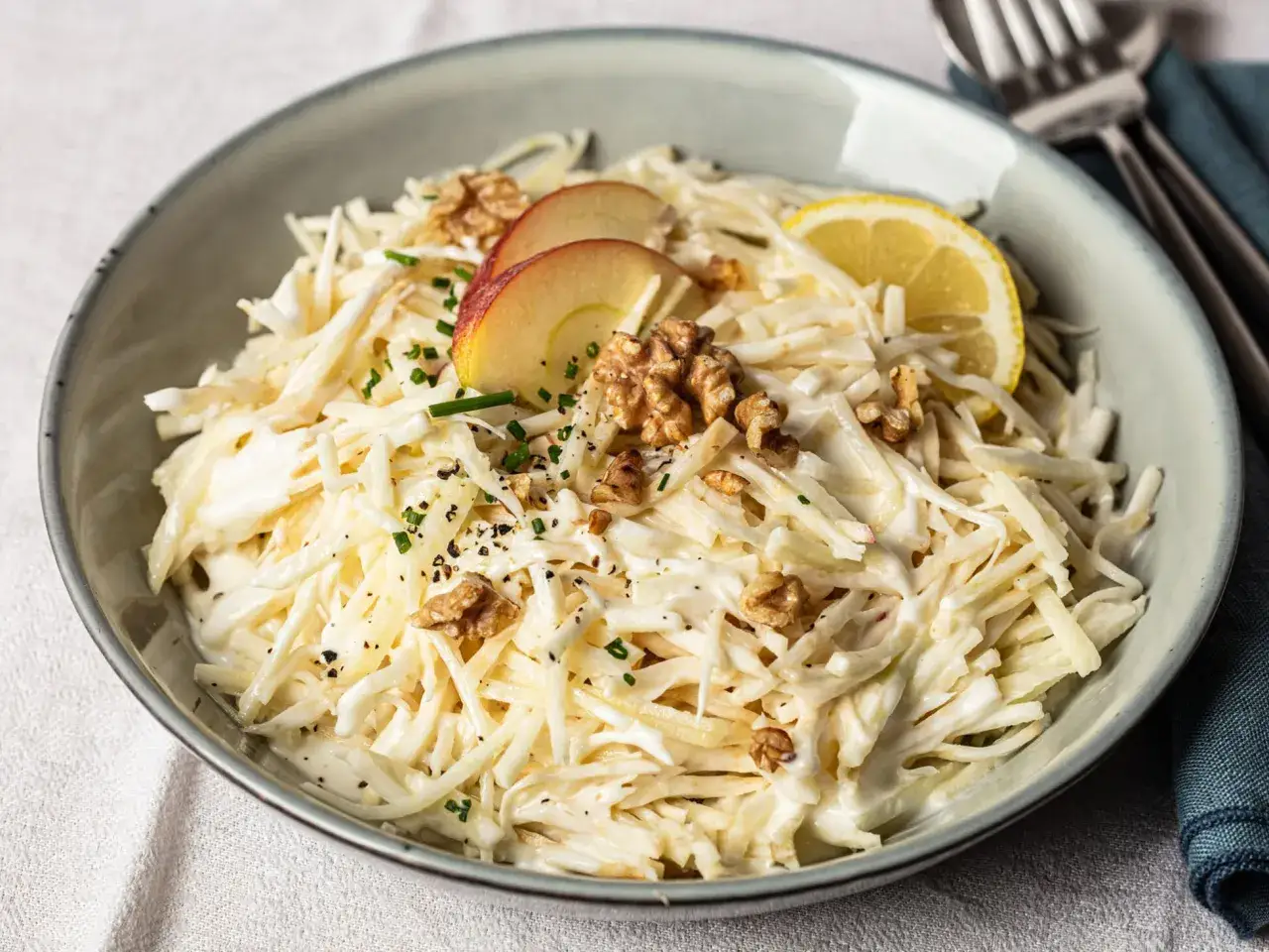 Ein cremiger Selleriesalat mit Äpfeln, Walnüssen und einem Zitronenschnitz, garniert mit Schnittlauch und Pfeffer.