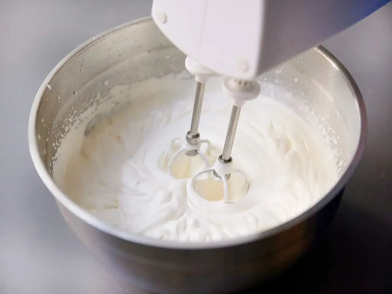 Ein Handmixer schl&auml;gt gerade Sahne in einer Edelstahlsch&uuml;ssel. Die aufgeschlagene Sahne ist cremig und volumin&ouml;s.