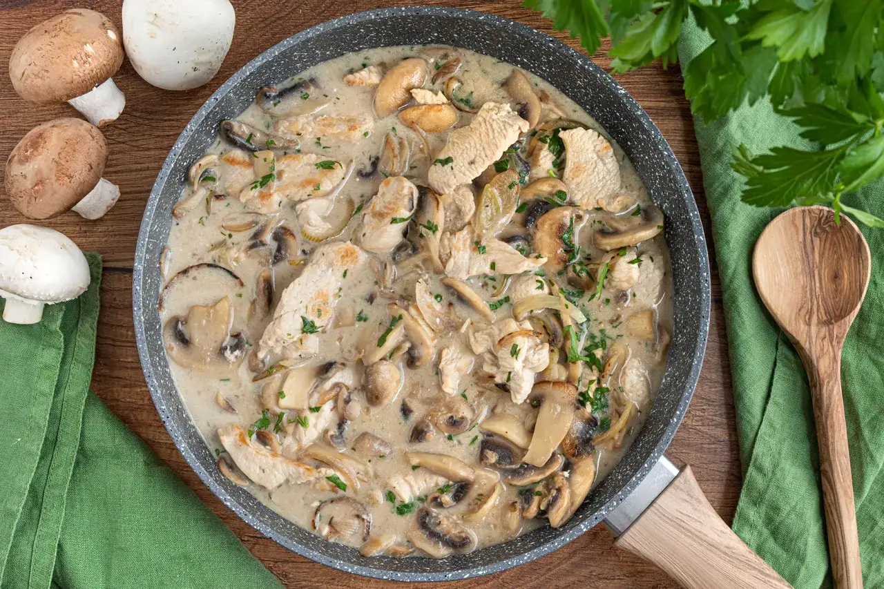 Herzhaftes Puten Geschnetzeltes mit Pilzen in cremiger So&szlig;e, garniert mit Petersilie. Daneben frische Champignons und ein Holzl&ouml;ffel.