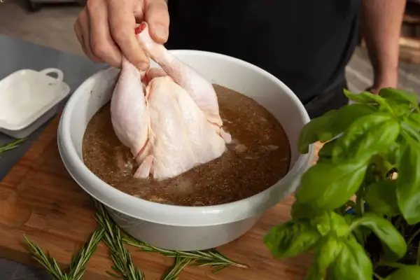 Ein rohes Hähnchen wird in eine Schüssel mit einer würzigen Flüssigkeit getaucht, um Fleisch in Salzlake einzulegen. Rosmarin und Basilikum liegen daneben.